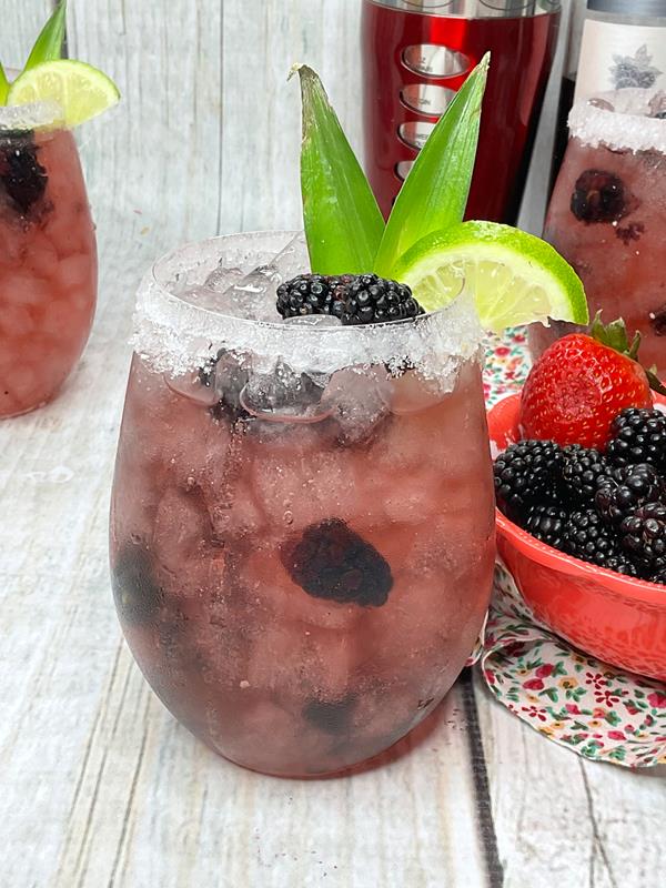 Close up of Rum Berry Cocktail, stemless wine glass filled with ice, blackberries and pinkish purple drink. Topped with blackberries, lime wedge, and pineapple heads. 