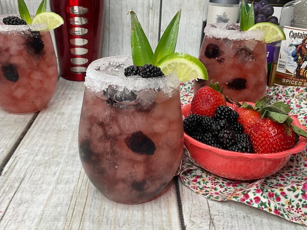 Three Rum Berry cocktails on a wooden table with a red bowl full of strawberries and blackberries. 