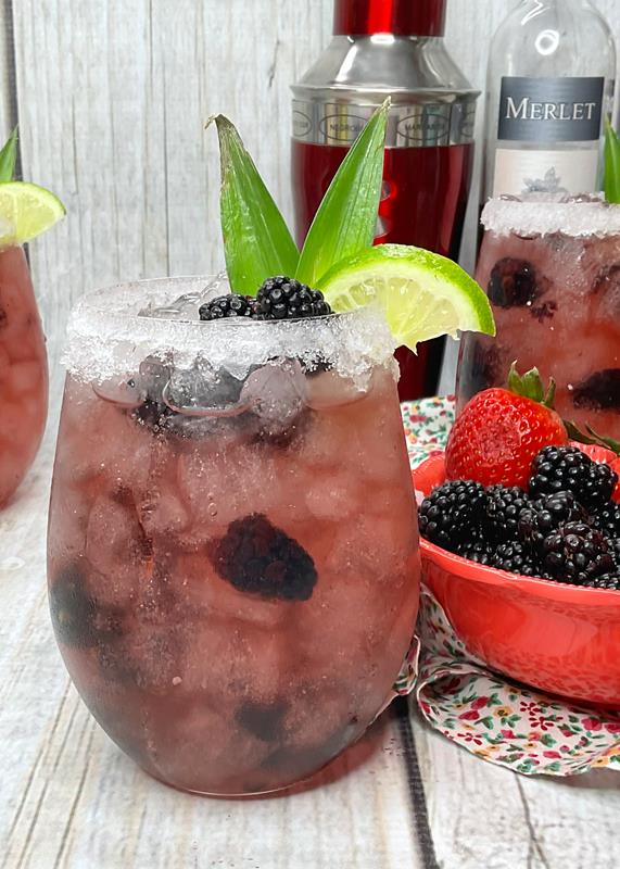 Cocktail with bottles of alcohol in background and red bowl of strawberry and blackberries in it. 