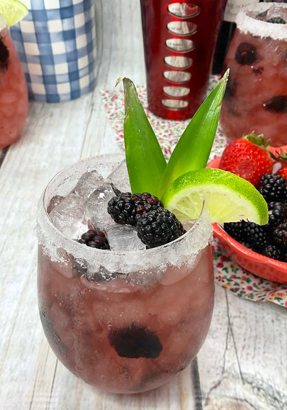 Slightly above view of cocktail, showing lots of ice, blackberries, pineapple heads, and lime wedge as garnish. 