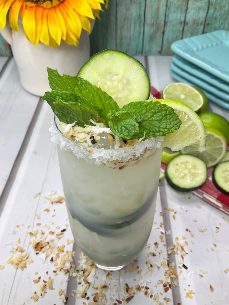 Slightly above view of Margarite showing toasted coconut on rim with salt. Mint leaves, cucumber and lime slices as garnish. 