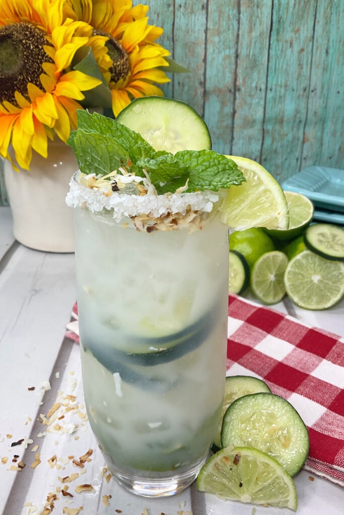Completed Margarita with coconut on rim, mint, cucumber, and lime slices as garnish. with sunflowers and limes in the background. 