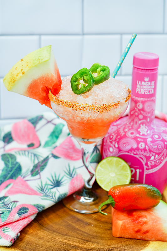 Pink frozen margarita drink with lime, jalapeno, watermelon and a bottle of tequila around it. 