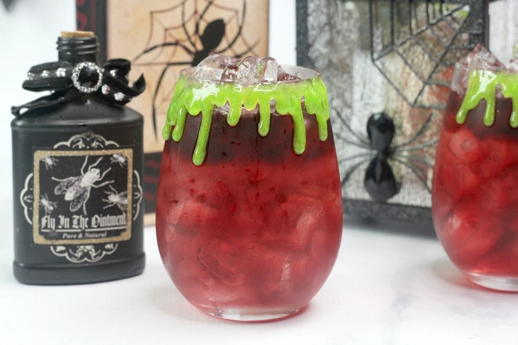 A red drink with ice in a rounded glass is topped with green "slime" that drips down the rim. The background features spooky Halloween decor, including a black potion bottle with a spider label and a framed spider web design.